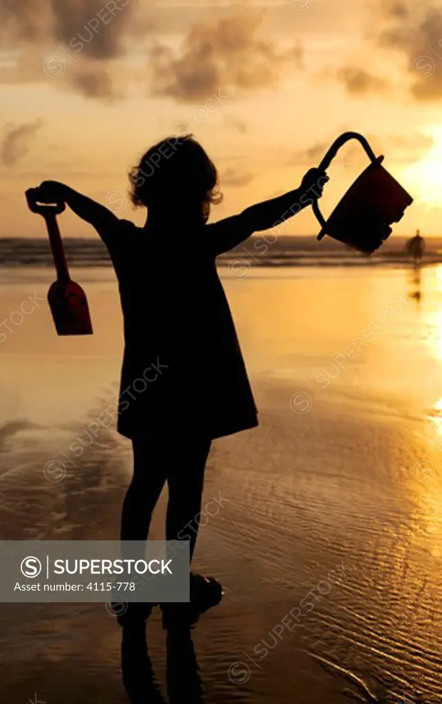 Little girl with bucket and spade silhouetted against setting sun on Summerleaze beach, Bude, North Cornwall, UK