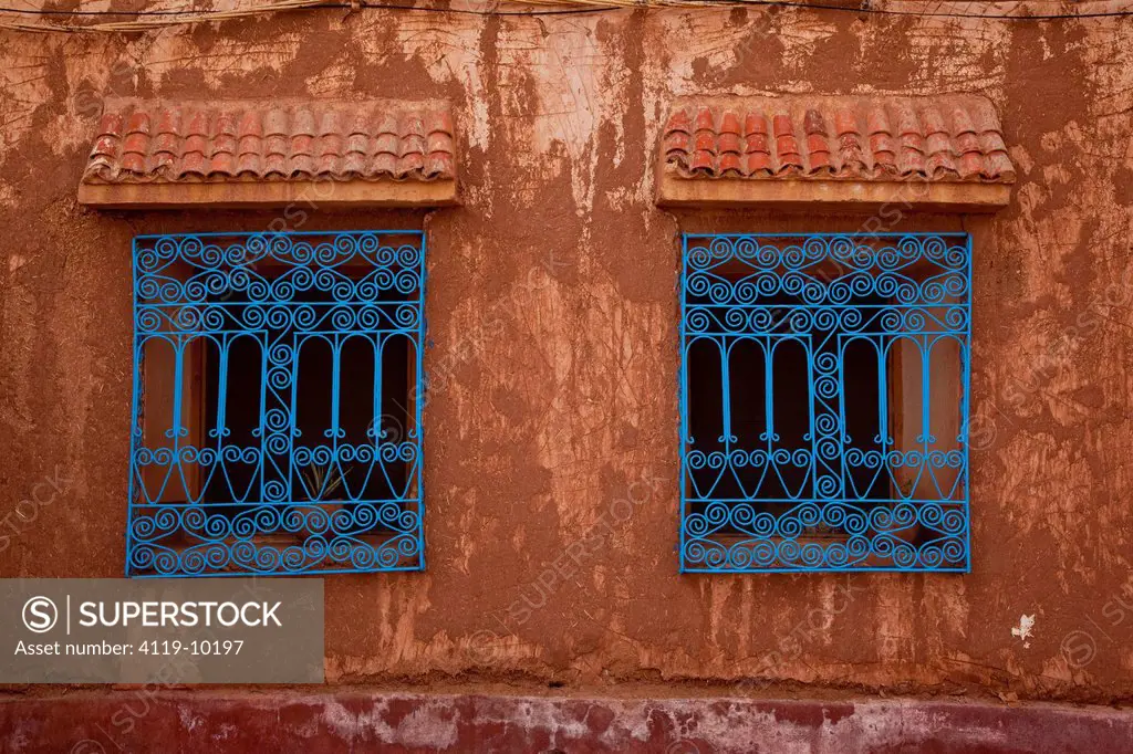 Photograph of a mud house with two windows and blue grilles on them in the village of Ait Benhaddou , Morocco