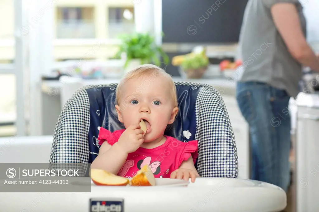 Girl sitting in baby-chair, eating some fruits. Her mother standing in the background.