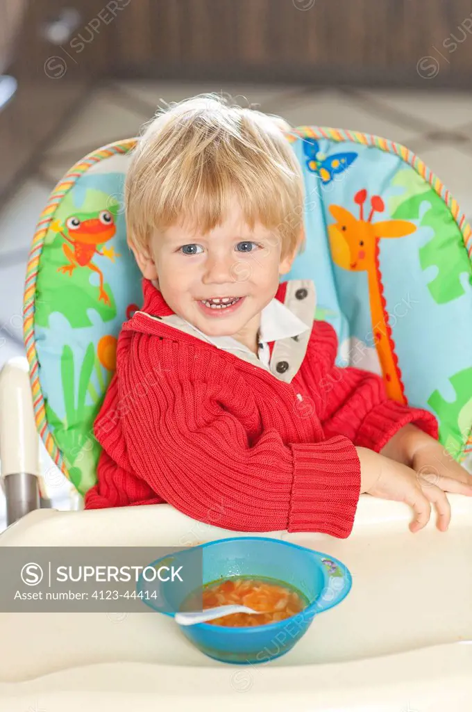 Adorable boy sitting in baby chair, eating soup.