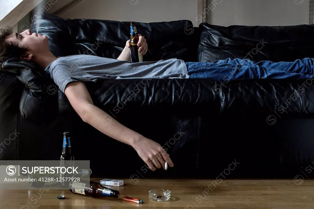 Young man lying on sofa holding beer and smoking cigarette.