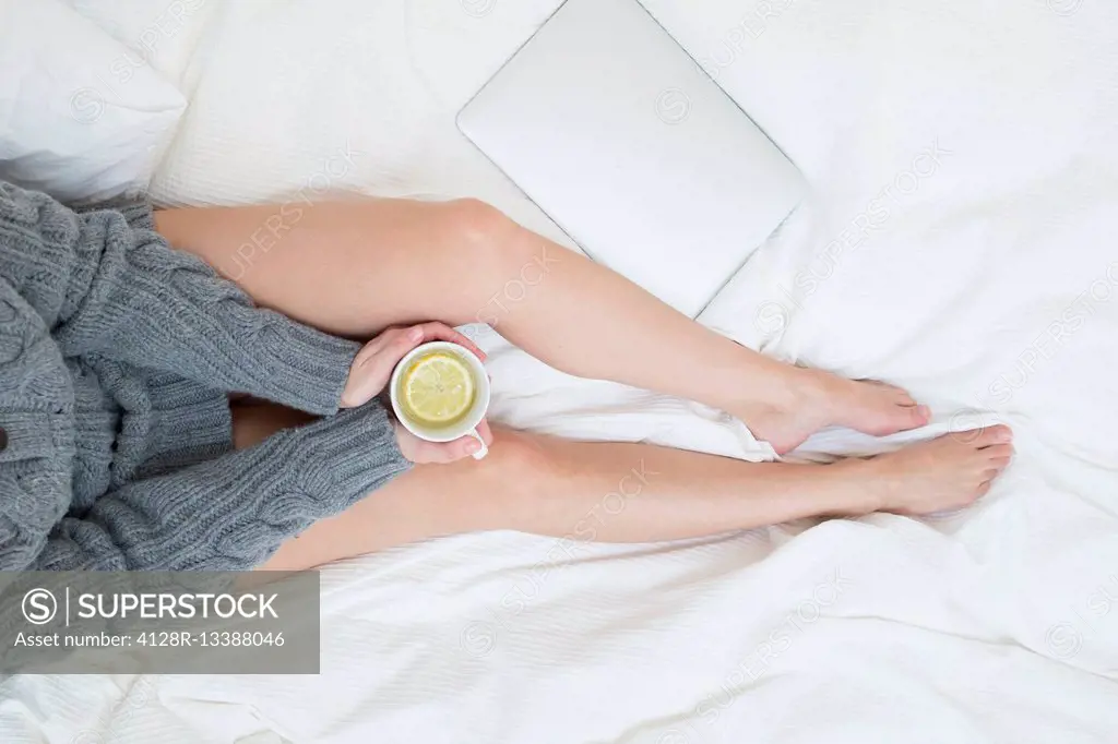 MODEL RELEASED. Woman sitting on bed with cup of hot lemon drink.