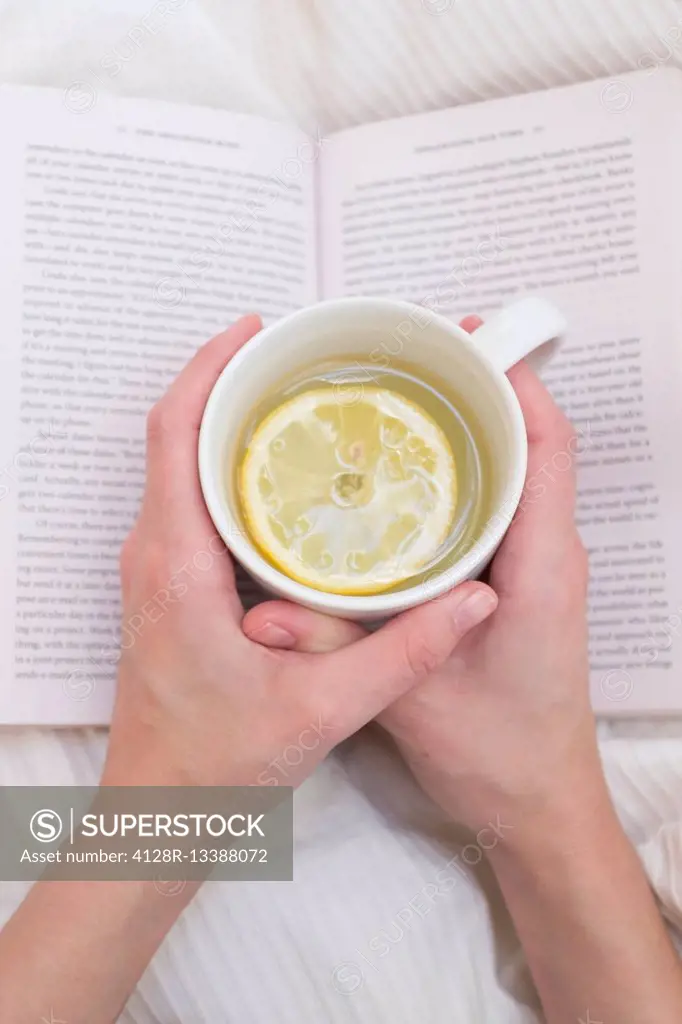 MODEL RELEASED. Woman holding cup of hot lemon drink and book.