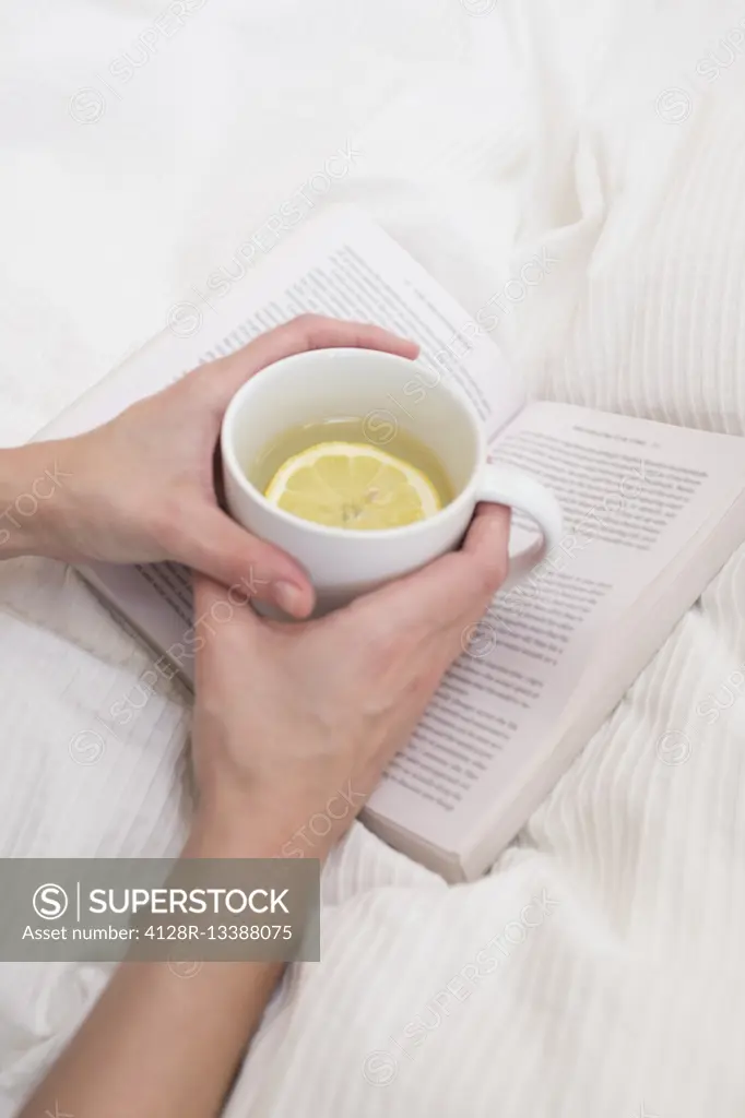 MODEL RELEASED. Woman holding cup of hot lemon drink and book.