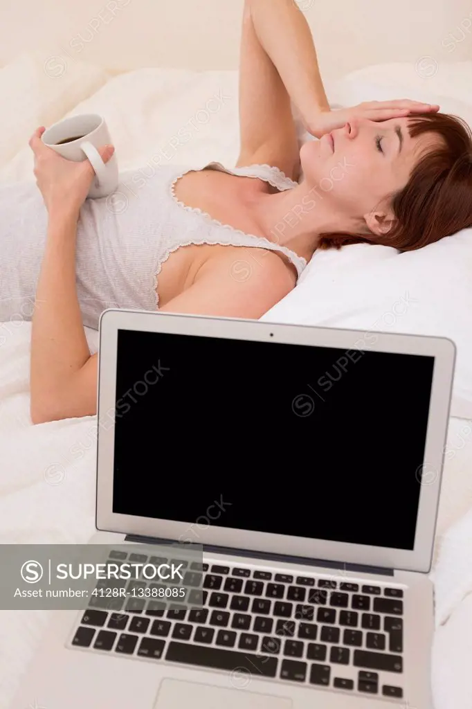 MODEL RELEASED. Exhausted woman in bed with laptop and black coffee.