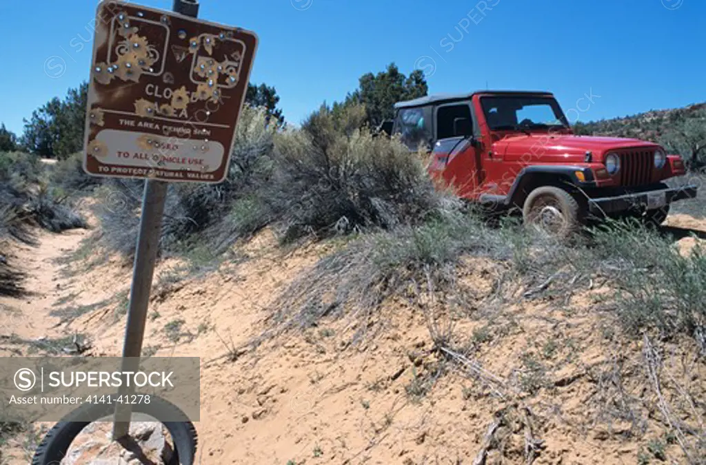 bullet riddled sign stating that the area is closed to all vehicle use. a jeep is parked nearby. off road rage