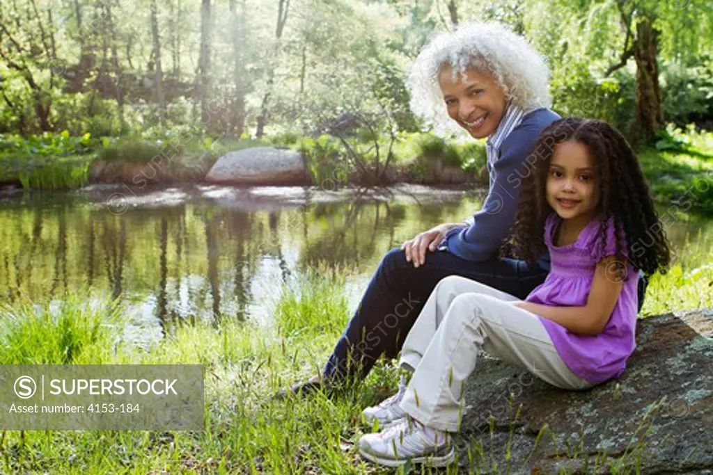 USA, Grandmother and granddaughter sitting on boulder near pond