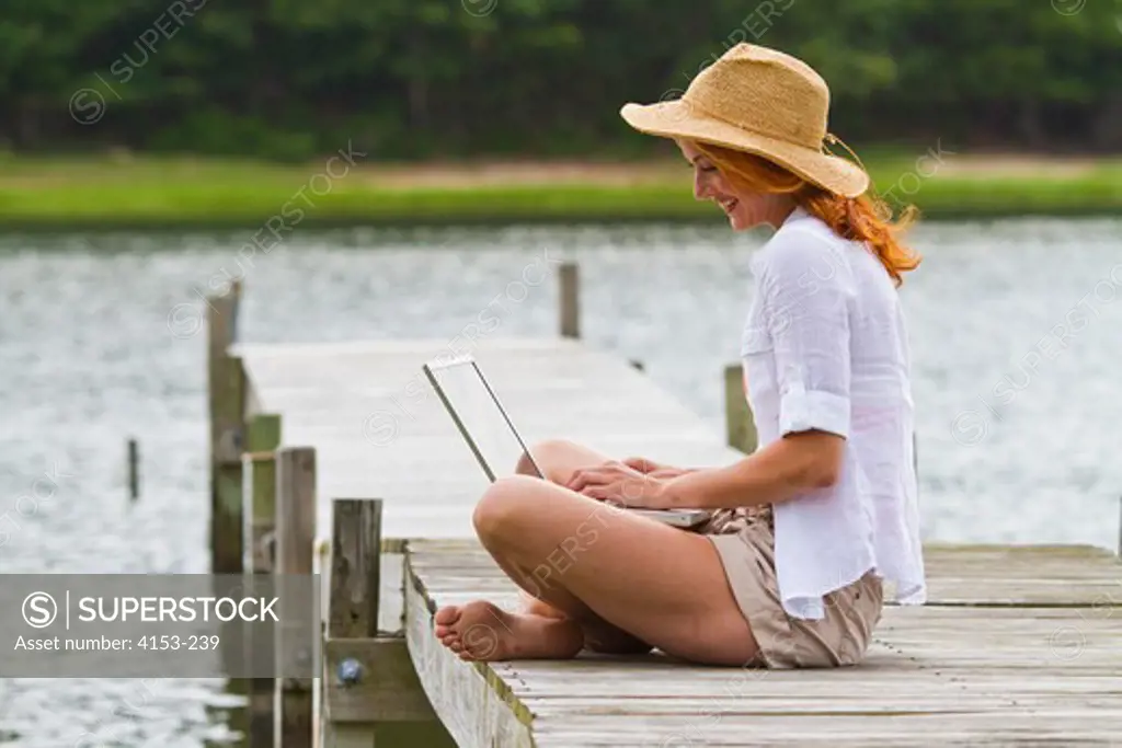 Young woman wearing straw hat using laptop on jetty
