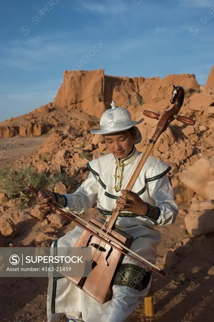 Mongolian Teenage Boy In Traditional Dress Performing Traditional Music With Horse Head Fiddle (Morin Khuur) At The Orange Rocks Of Bayan Zag, Commonly Known As The Flaming Cliffs In The Gobi Desert, Mongolia