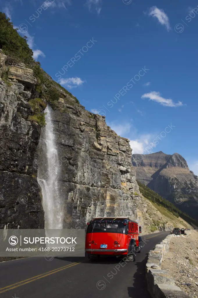 Red tour bus at waterfall coming down next to the Going-to-the-Sun Road near Logan Pass in Glacier National Park, Montana, United States.