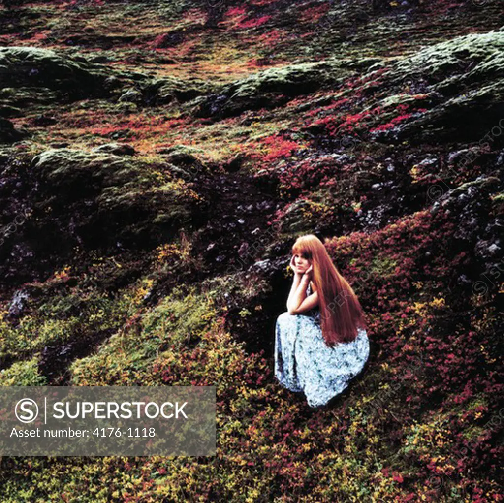 Woman with long hair wearing a dress, sitting in a mossy lava field, resting head in hands, Iceland