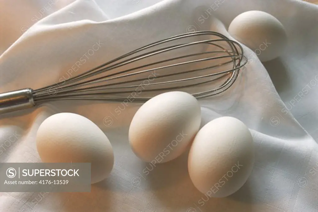 Still life of whisk with chicken eggs on white cloth in kitchen