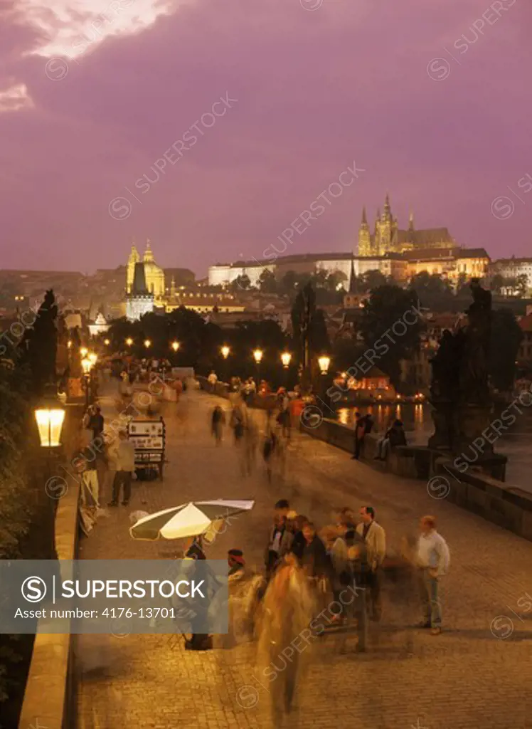 Artists and tourists on Charles Bridge below street lamps and Prague Castle at night