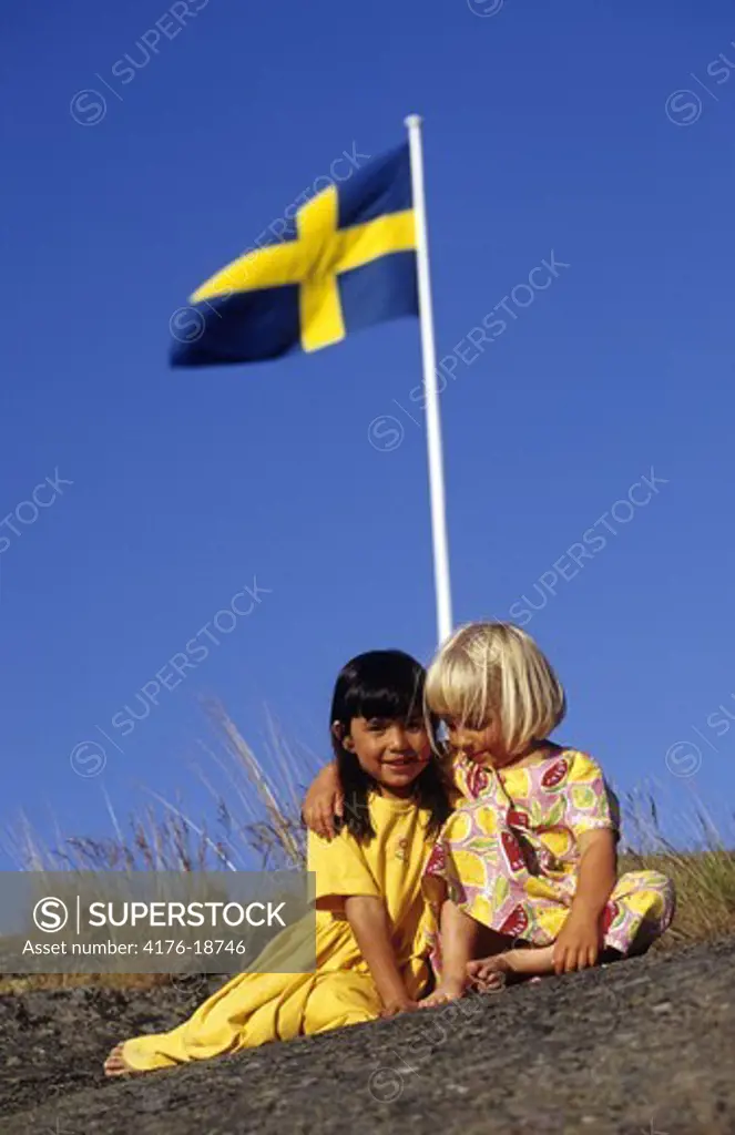 Two girls sitting by a swedish flag against blue sky