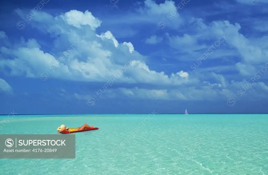 Woman floating on air mattress and aqua waters under white clouds with sailboat on distant horizon
