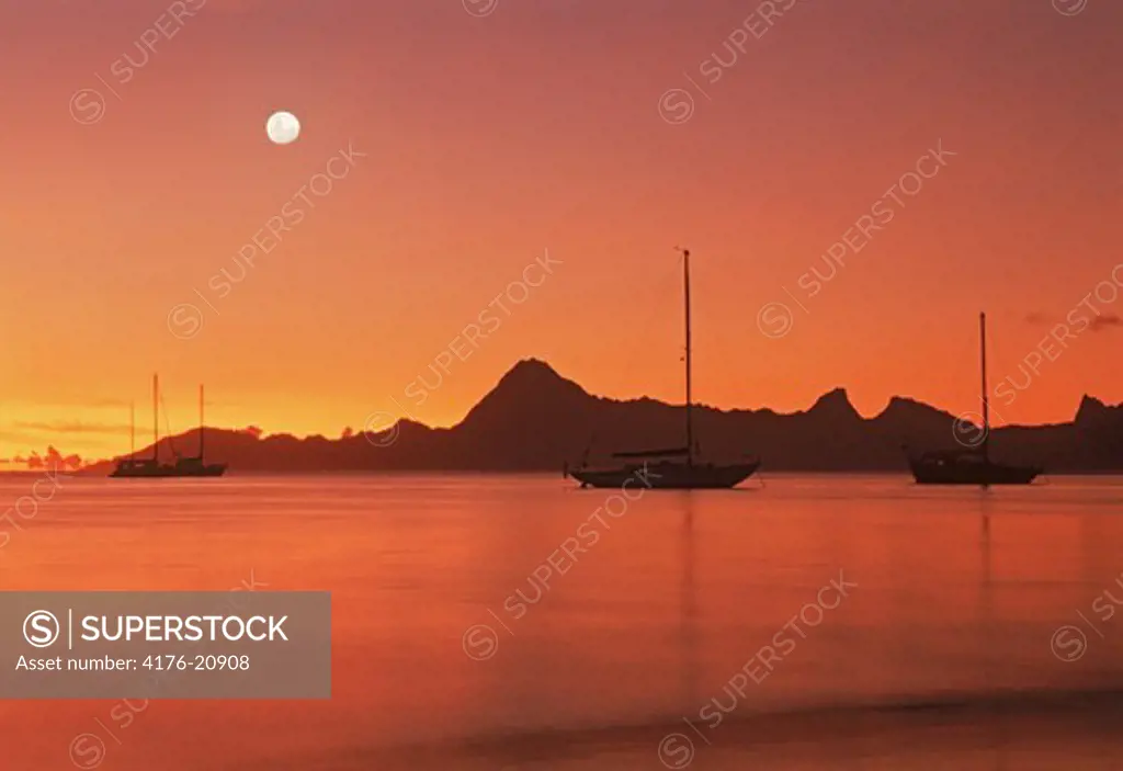Full moon over distant Moorea from island of  Tahiti with sailboats anchored offshore at sunset