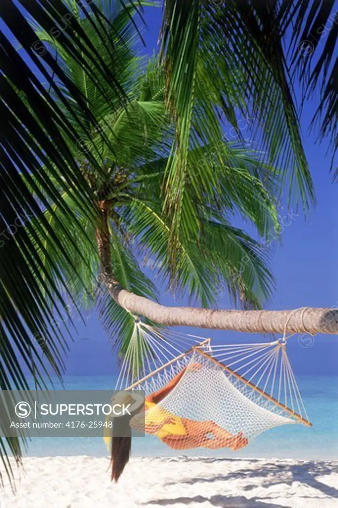 Woman with hat and yellow bathing suit relaxing in hammock under green palms over white sandy beach