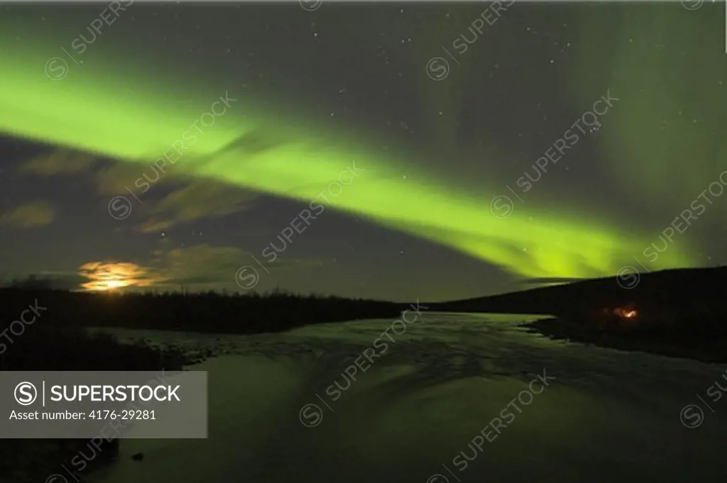 Norther lights and moon over open waters and campfire by Lainio River Swedish Arctic tundra. Lapland. Sweden