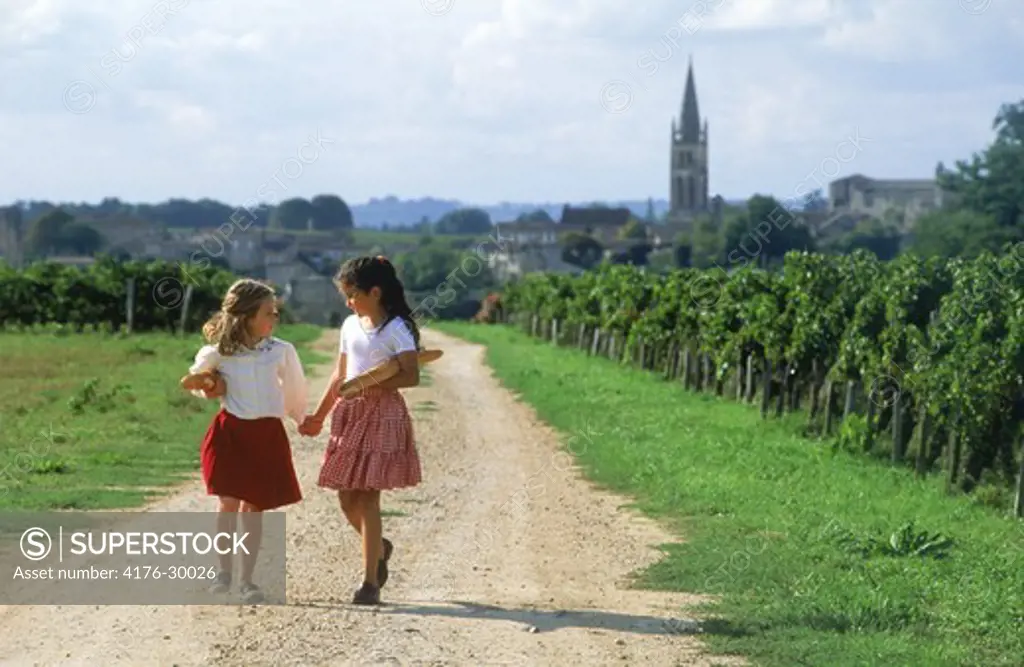 Two school girls walking through vineyards with baguettes near village of St Emilion in France