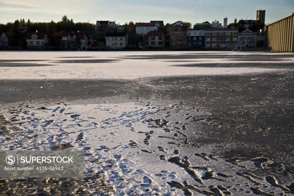 Footprints on a frozen lake (Tjornin). Houses in the background. Downtown Reykjavik. Iceland