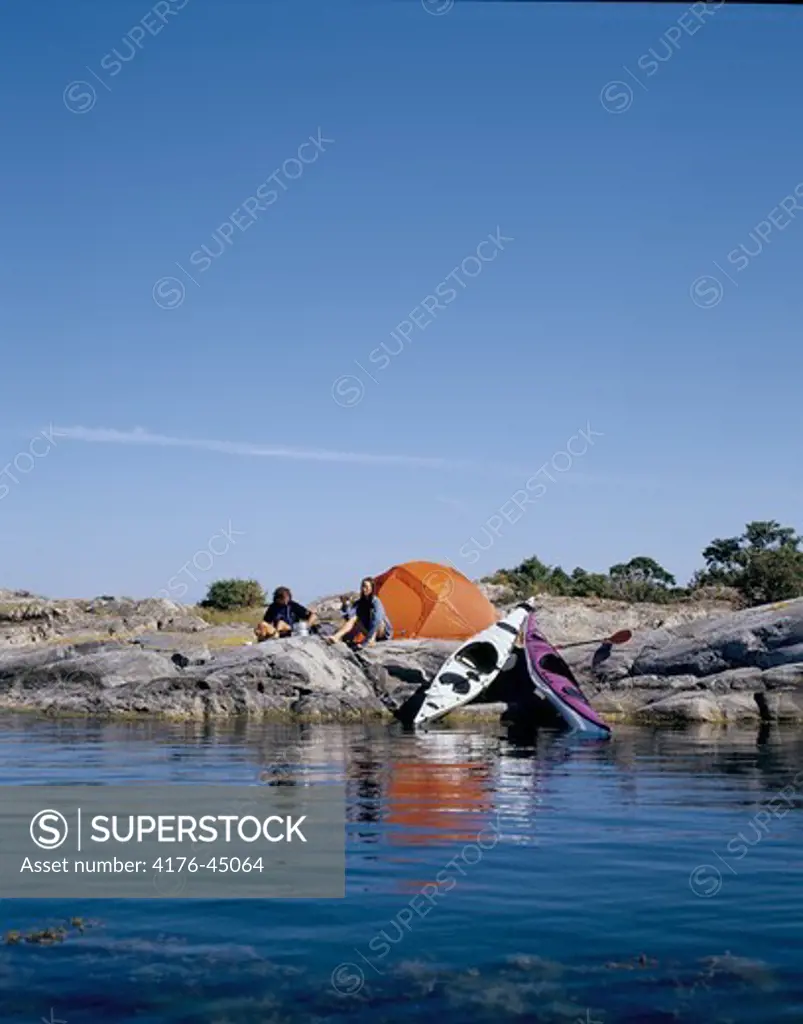Two people sitting near the tent with kayaks on the rocky beach
