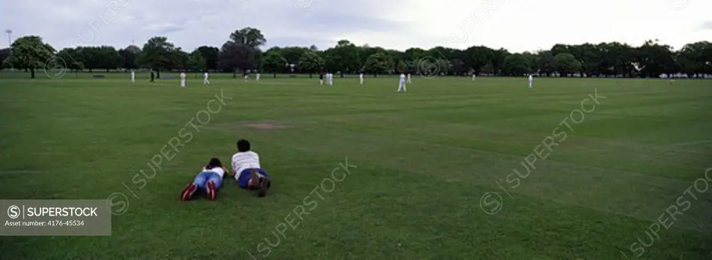 Two people lying watching a cricket game