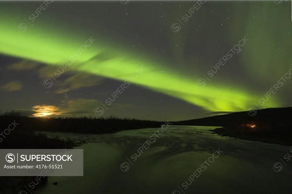 Northern lights and moon over open water and campfire by Lainio River Swedish Arctic tundra, Lapland
