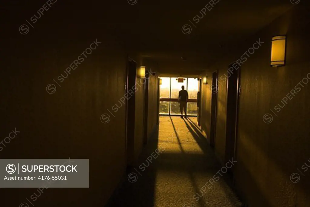 A man standing by a window at the end of a corridor, watching the rising sun, Miami, USA.