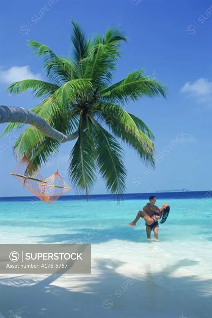 Man carrying woman out of aqua colored waters under plam tree