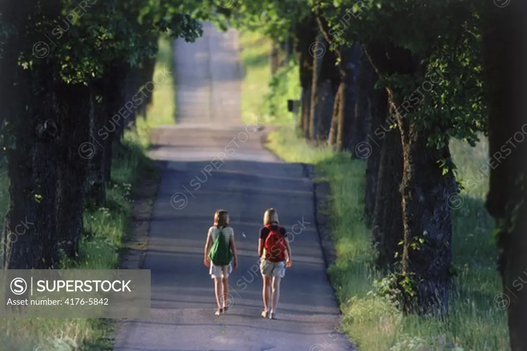 Two young girls with school backpacks walking on tree-lined country road in Sweden