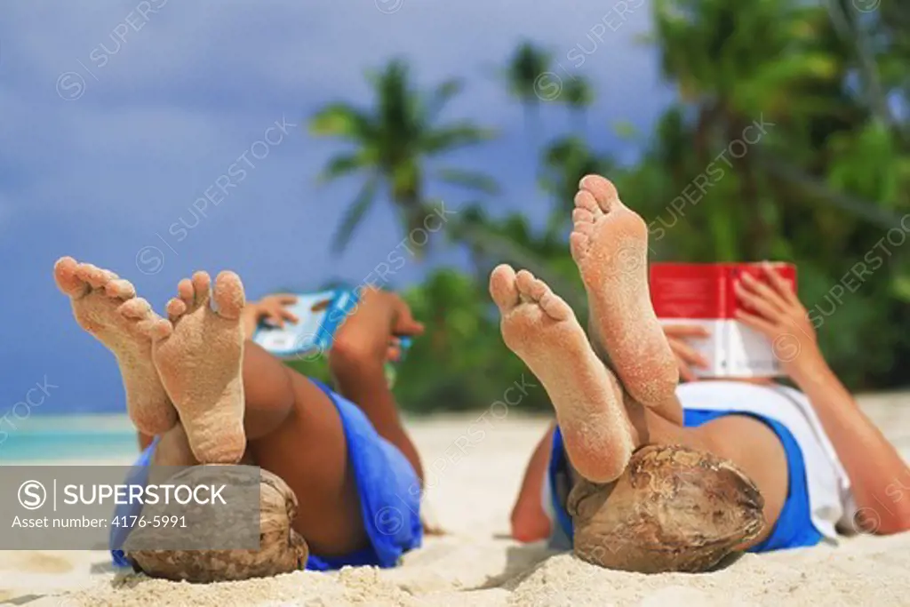 Couple reading books on sandy beach with feet resting on coconuts