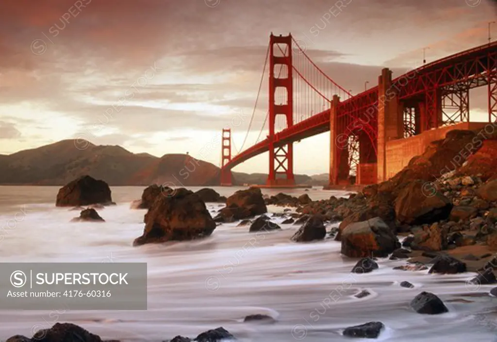 Waves hitting rocky shoreline along Baker Beach below Golden Gate Bridge in San Francisco at sunset