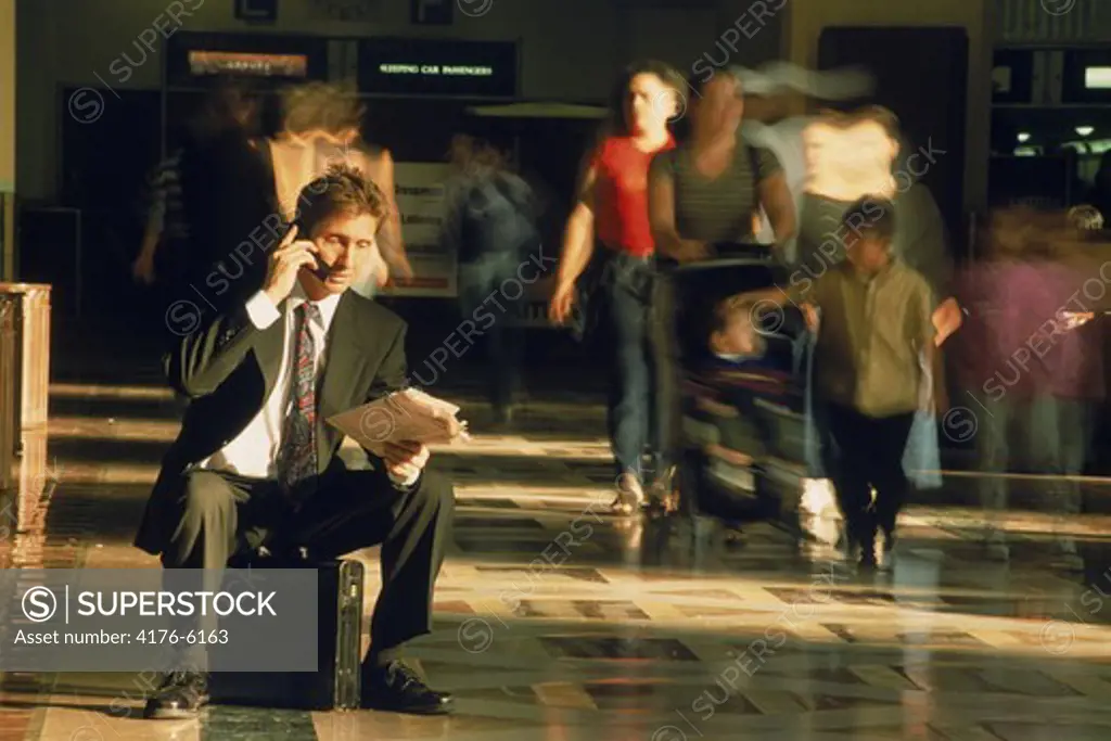 Businessman sitting on briefcase talking on cellphone and reading paper in Los Angeles Train Station