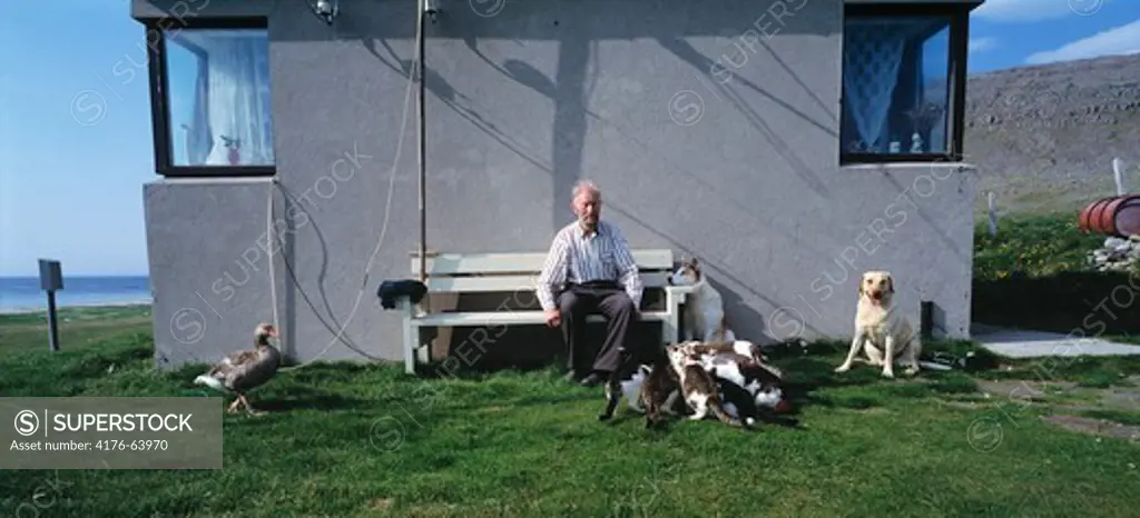 Senior man sitting infront of his house in Hvallatur with group of animals infront of it