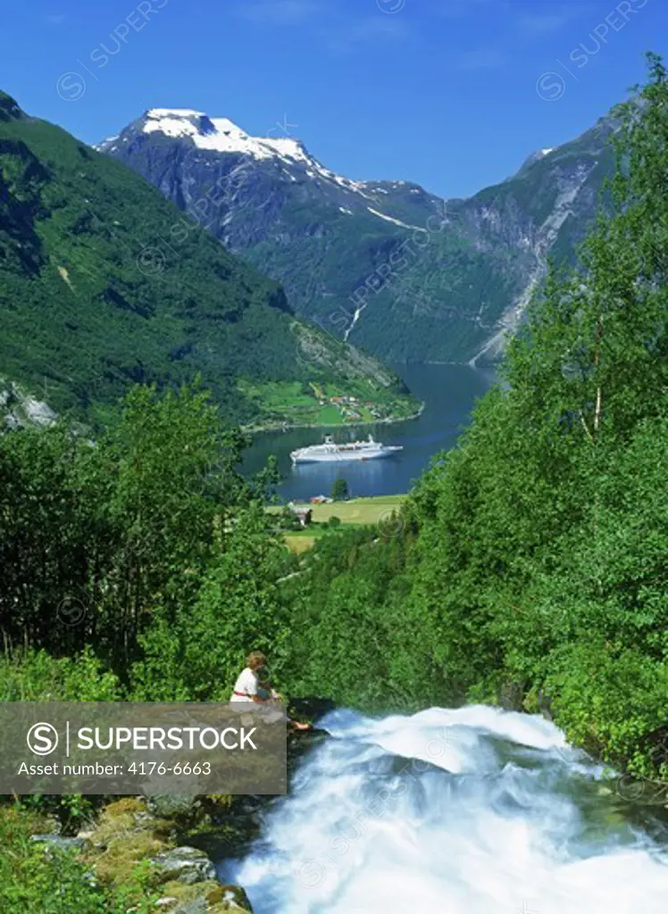 Woman next to stream dropping into Geirangerfjord with passenger ship