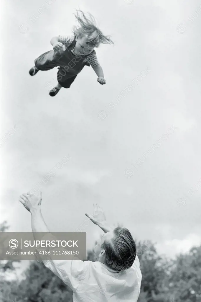 1950s 1960s FUN OR DANGEROUSLY FOOLISH FATHER THROWING TODDLER DAUGHTER HIGH INTO THE AIR AND WAITING TO CATCH HER COMING DOWN