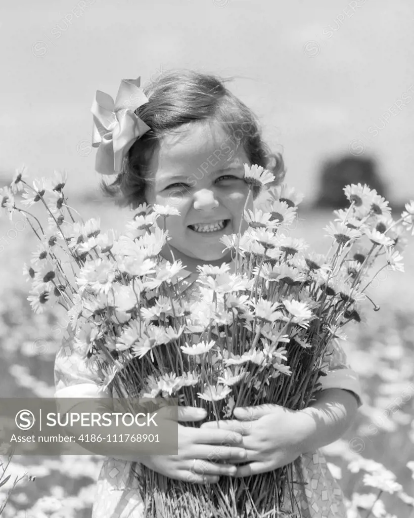 1930s SMILING HAPPY LITTLE GIRL LOOKING AT CAMERA HOLDING A BIG BUNCH OF FRESHLY PICKED DAISIES FLOWERS