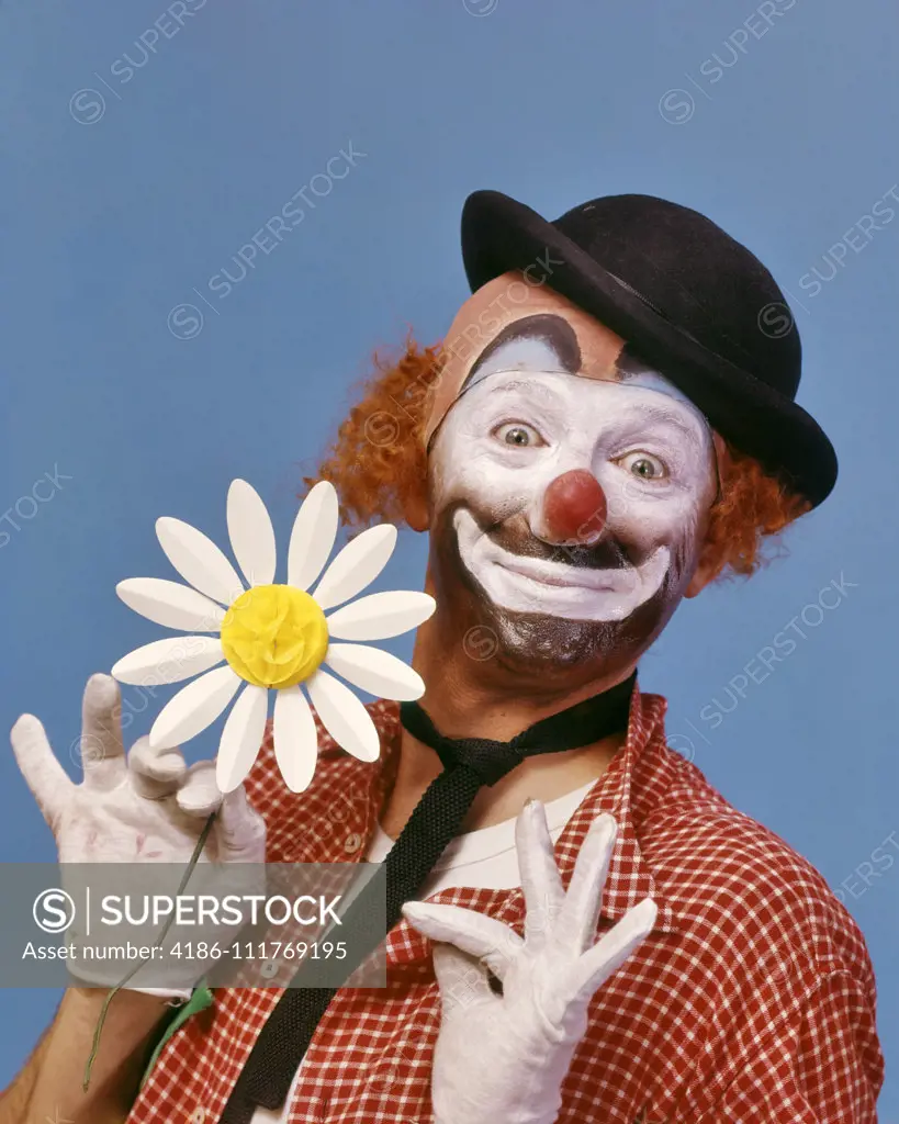 1970s SMILING WIDE-EYED WHITE FACE HOBO CLOWN WITH RED NOSE LOOKING AT CAMERA WEARING A DERBY HAT AND HOLDING A PAPER DAISY