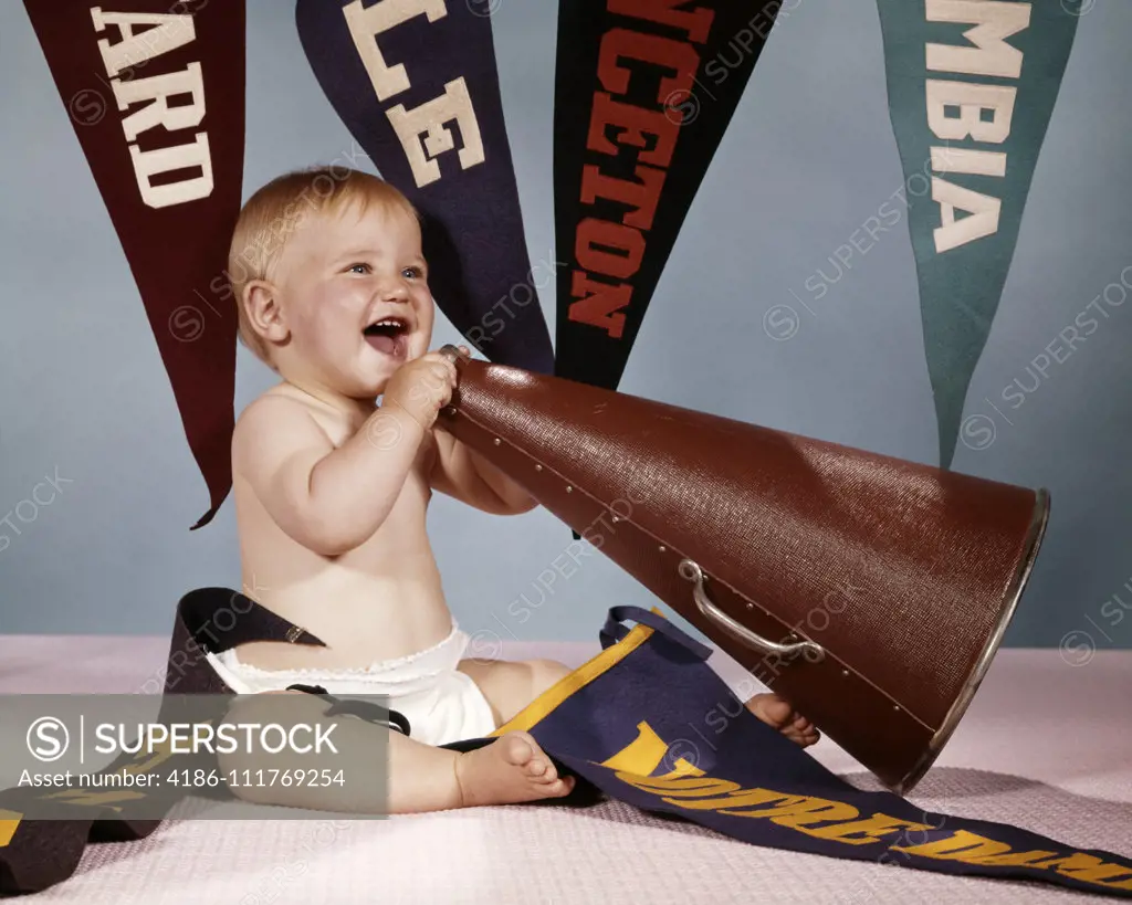 1960s EXCITED BABY GIRL CHEERLEADER SHOUTING INTO MEGAPHONE SURROUNDED BY COLLEGIATE ATHLETIC PENNANTS