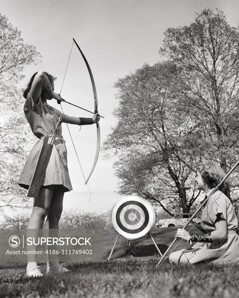 1950s TWO ANONYMOUS TEENAGE GIRLS PRACTICING ARCHERY TOGETHER AT CAMP IN COUNTRY