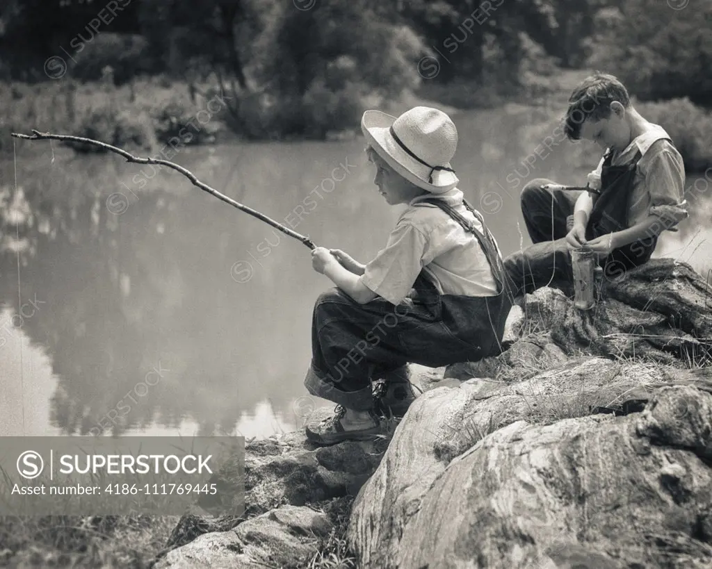 1930s  TWO BOYS  SITTING ON ROCKS FISHING IN POND WITH POLES MADE FROM TWIGS AND STRING