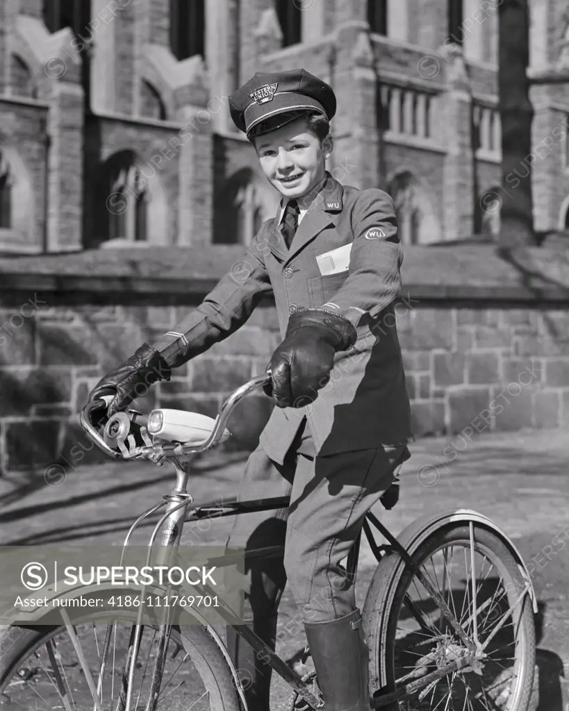 1940s SMILING WESTERN UNION TELEGRAM DELIVERY TEEN BOY IN UNIFORM WEARING LEATHER GLOVES LOOKING AT CAMERA RIDING ON BICYCLE