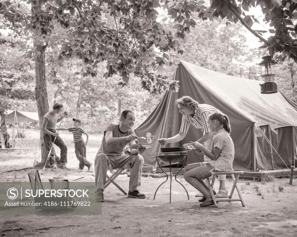 1950s 1960s FAMILY OF 5 TENT CAMPSITE MOM COOKING BREAKFAST SERVING DAUGHTER AND DAD TWO BOYS LEANING ON TREE WITH FISHING ROD
