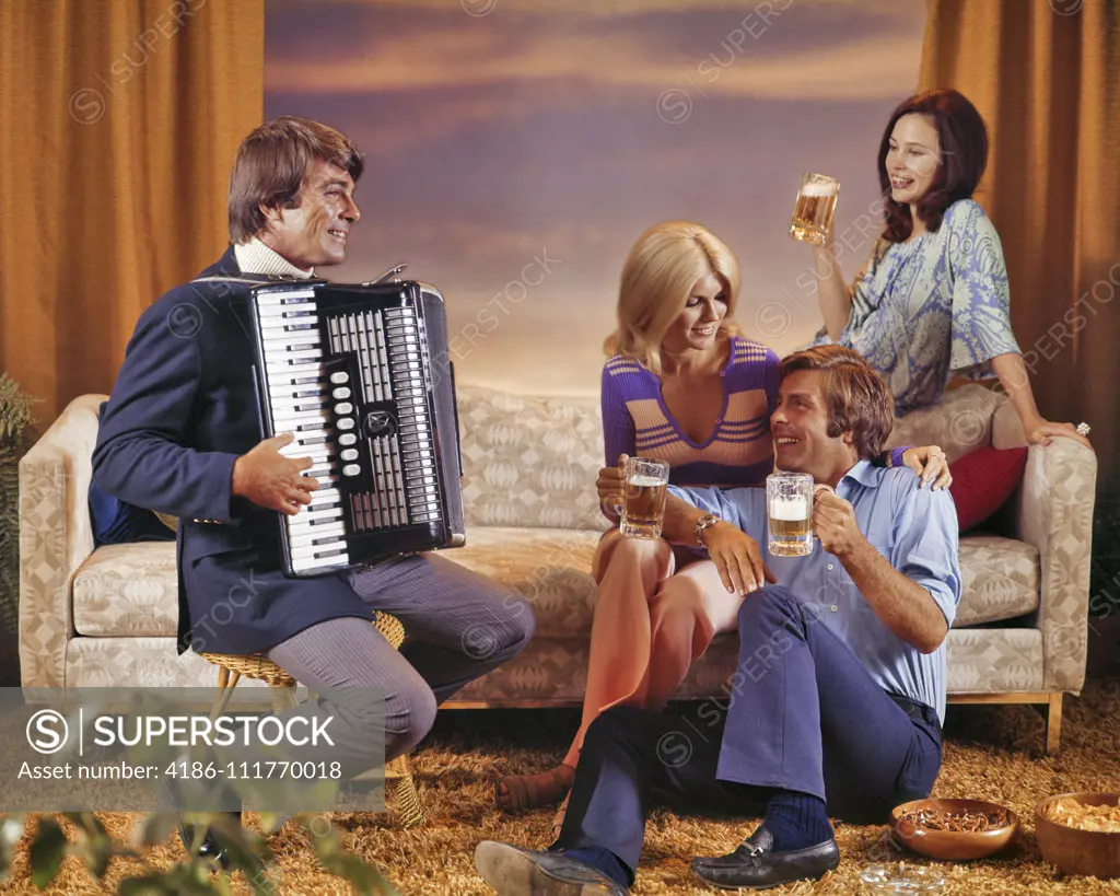 1970s 1980s TWO SMILING COUPLES MEN AND WOMEN INFORMALLY SOCIALIZING THREE DRINKING BEER AND ONE MAN PLAYING AN ACCORDION