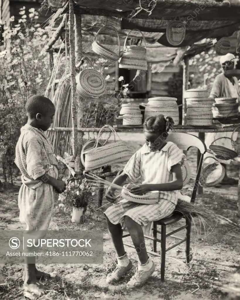 1940s AFRICAN AMERICAN BOY STANDING WATCHING GIRL WEAVING BASKET AT STAND WITH SEVERAL BASKETS FOR SALE SOUTH CAROLINA USA
