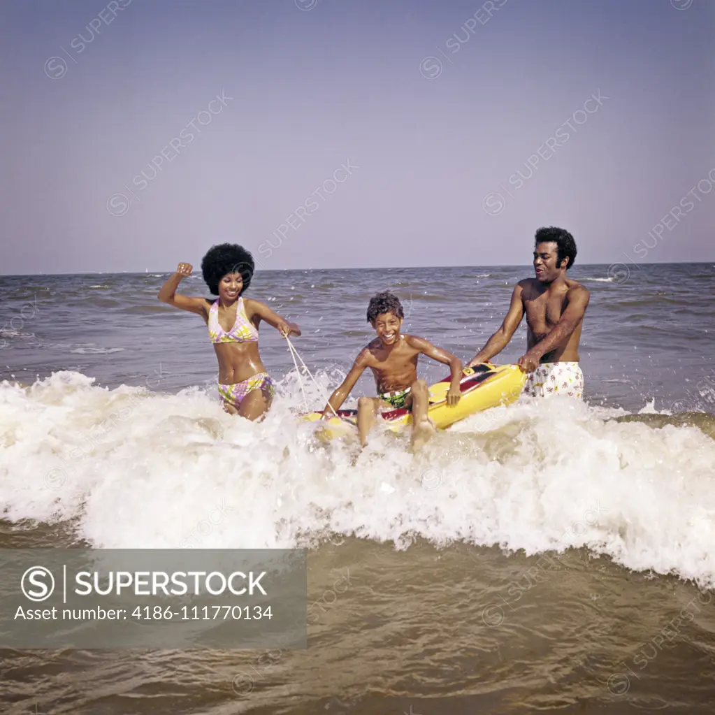 1970s AFRICAN-AMERICAN FAMILY JUMPING IN OCEAN SEASHORE SURF MOTHER AND FATHER WITH SON ON INFLATABLE RAFT