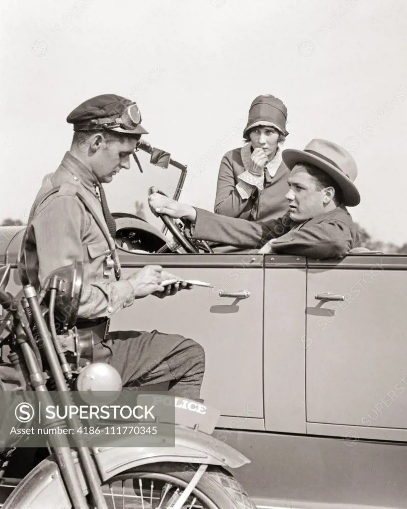 1920s MOTORCYCLE POLICEMAN WRITING A SPEEDING TICKET TO A COUPLE SITTING IN CONVERTIBLE SEDAN