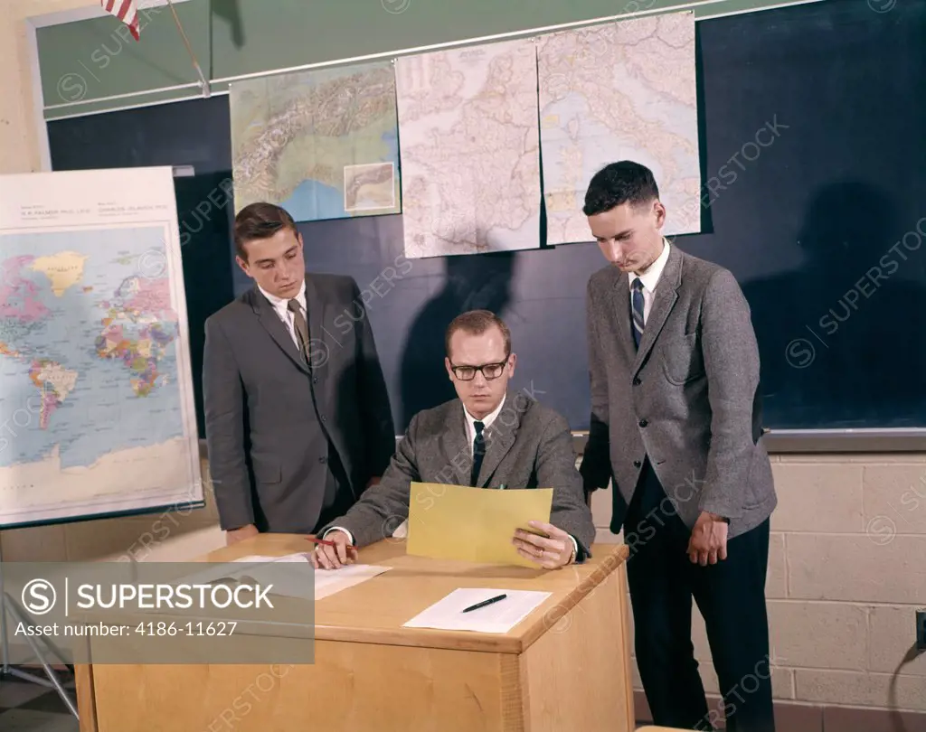 1960S Teacher Sitting At Desk With Boy Students Looking At And Grading Paper Indoor 