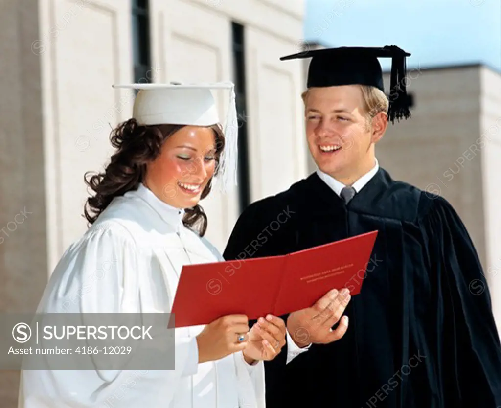 1970S Teen Couple Girl White Robe Boy In Black Robe Mortarboard Hold Red Diploma Between Them High School Smiling Proud