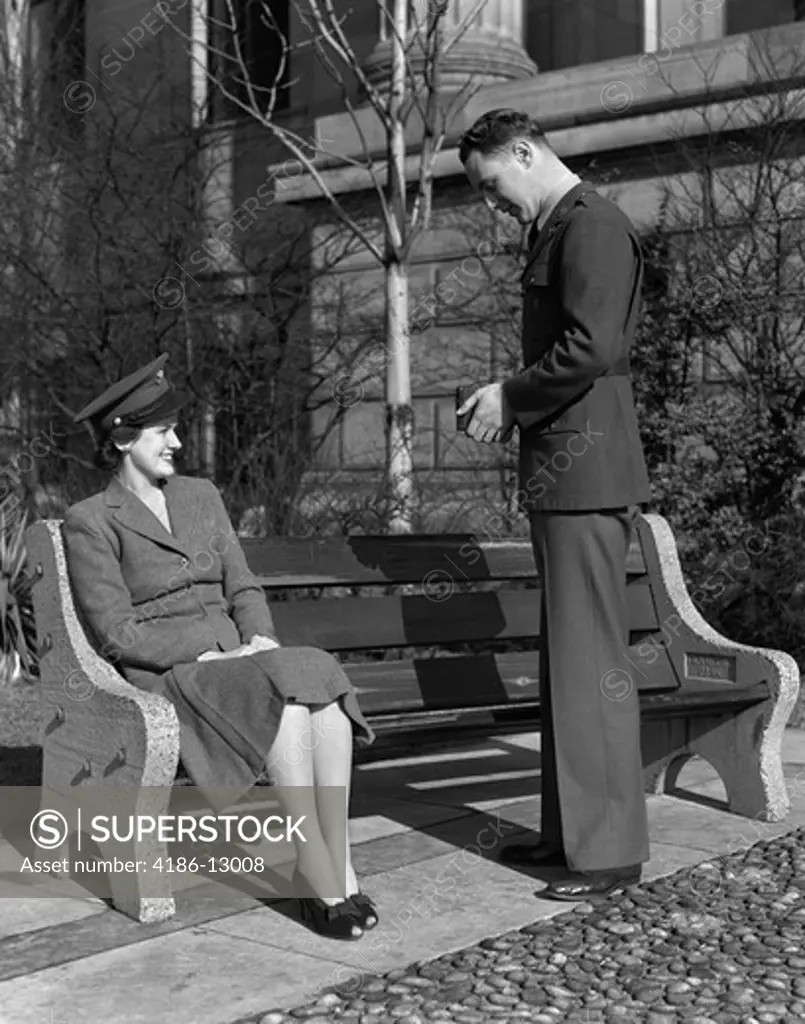 1940S Couple Man In Army Uniform Taking Photo Of Woman Sitting On Bench Wearing Hat